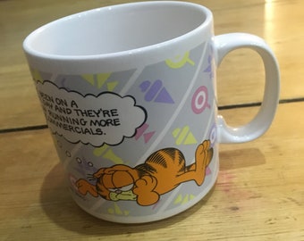 May include: White ceramic mug with a cartoon image of Garfield the cat sleeping. The mug has a gray and white geometric pattern with yellow, pink, and purple accents. The text on the mug reads "Been on a day and they're running more commercials."