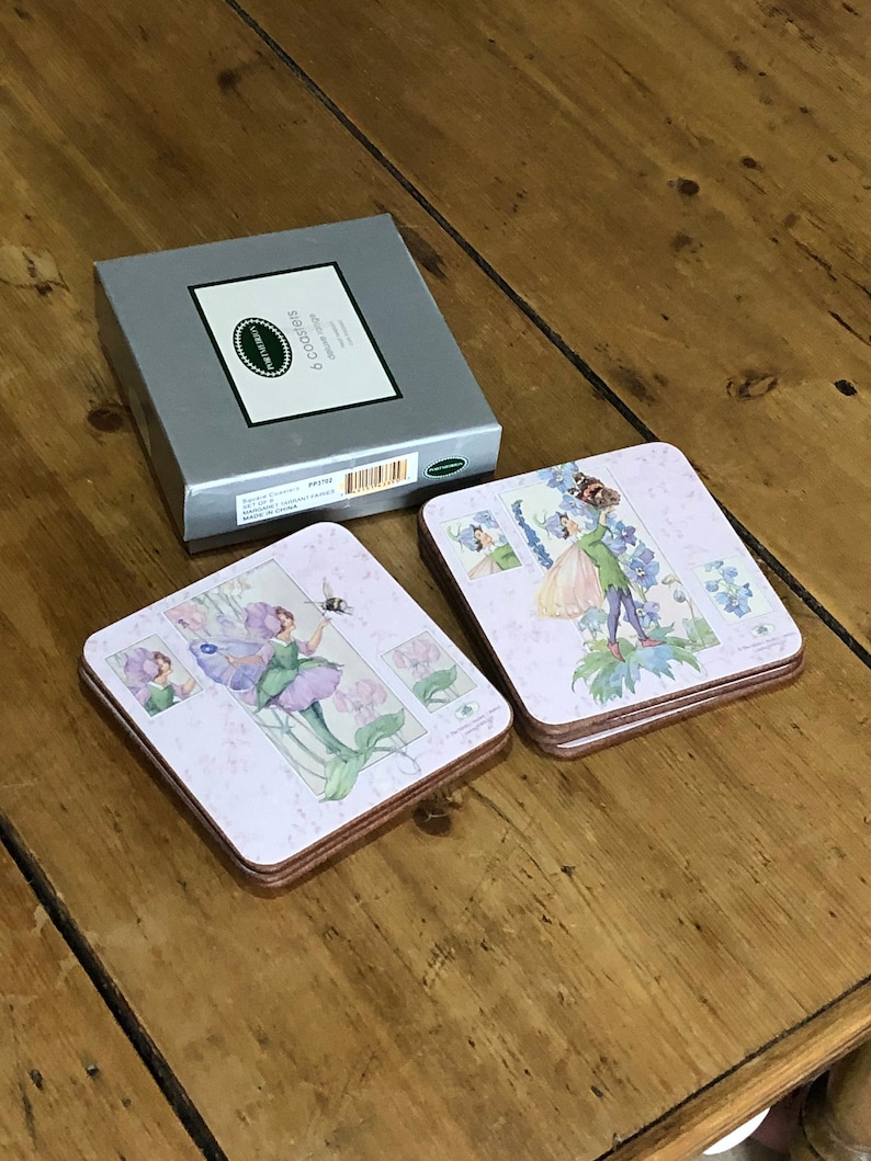 May include: A set of six coasters featuring a fairy design with purple flowers and a bee. The coasters are made of cork and have a pink background. The box for the coasters is silver with the text "6 Coasters" printed on it.