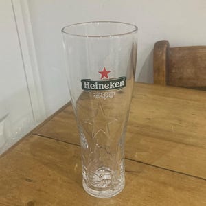 Op de afbeelding: Een helder, hoog bierglas met het Heineken-logo in groen en rood. Het glas heeft een sterontwerp dat in het glas is ge&euml;tst. Het glas staat op een houten tafel.