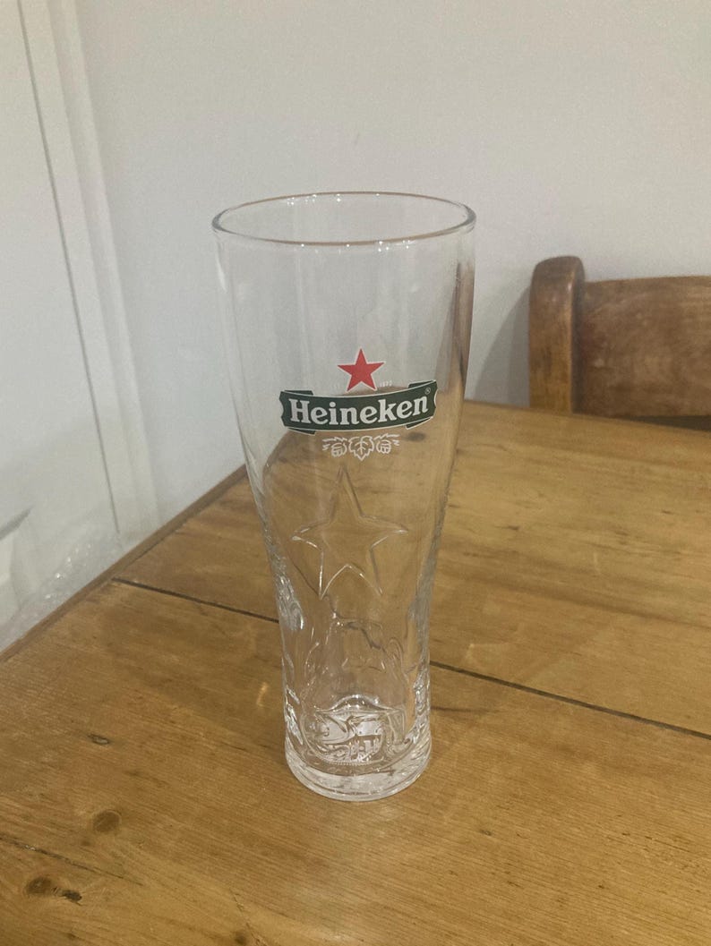 Op de afbeelding: Een helder, hoog bierglas met het Heineken-logo in groen en rood. Het glas heeft een sterontwerp dat in het glas is ge&euml;tst. Het glas staat op een houten tafel.
