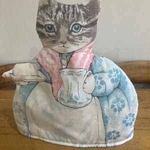 May include: A fabric tea cozy shaped like a cat, with a gray and white tabby pattern on the head. The cat wears a pink collar and a blue floral patterned dress, holding a tray and a teacup.