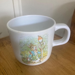 May include: White ceramic mug with a colourful illustration of Peter Rabbit holding carrots, and a robin perched on a shovel. The mug has a curved handle and is set on a wooden surface. The design is reminiscent of a children's book.