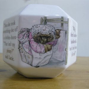 May include: A white ceramic piggy bank shaped like a hexagon with a Beatrix Potter illustration of a hedgehog wearing a pink dress and a white bonnet. The text "Then she took something else off a clothes-horse- "That isn't my pinny?" said Lucie." is printed on the side of the bank.
