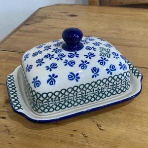 Könnte beinhalten: Eine Keramik-Butterdose mit Deckel, mit weißer Basis, verziert mit blauen Blumenmustern. Der Deckel hat einen dunkelblauen Knopf. Der Rand der Schale ist mit einem grün-blauen geometrischen Design verziert. Die Schale ist rechteckig mit abgerundeten Kanten.