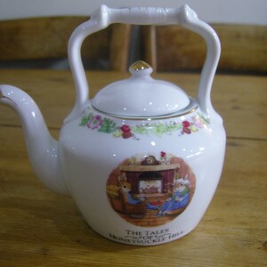 Pode incluir: Bule de porcelana branca com um design floral e uma cena de dois coelhos perto de uma lareira. A chaleira tem uma borda dourada e o texto "The Tales of Honeysuckle Hill".