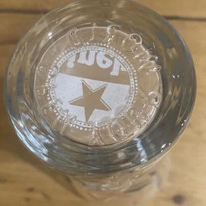 Op de afbeelding: Helder glas met een cirkelvormig ontwerp aan de onderkant. Het ontwerp toont een ster en het woord "Just" in een wit en goud kleurenschema. Het glas is waarschijnlijk een drinkglas of een vergelijkbare container.