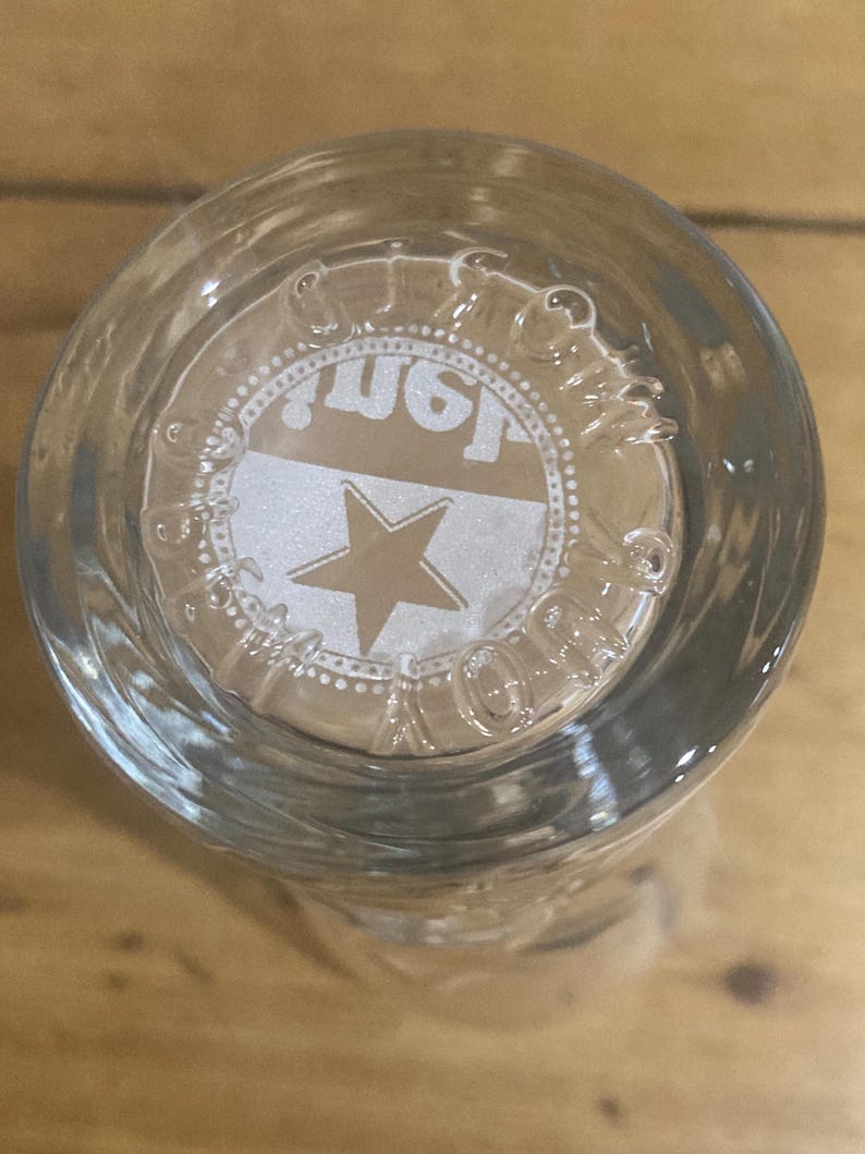 Op de afbeelding: Helder glas met een cirkelvormig ontwerp aan de onderkant. Het ontwerp toont een ster en het woord "Just" in een wit en goud kleurenschema. Het glas is waarschijnlijk een drinkglas of een vergelijkbare container.