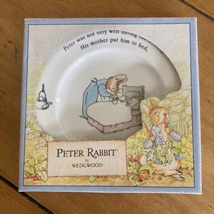 May include: A white ceramic plate featuring a Peter Rabbit illustration in a square box. The plate shows a rabbit in bed with text reading, "Peter was not very well during the night. His mother put him to bed." The box displays the Peter Rabbit by Wedgwood logo.
