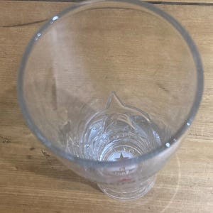 Op de afbeelding: Transparant bierglas met een stervormig ontwerp aan de basis. Het glas heeft een brede opening en een licht uitlopende basis. Het glas is leeg en staat op een houten oppervlak.