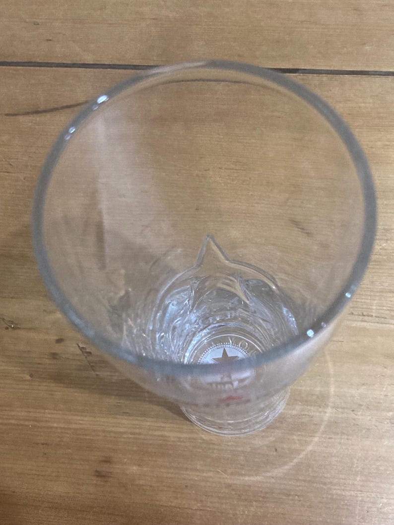 Op de afbeelding: Transparant bierglas met een stervormig ontwerp aan de basis. Het glas heeft een brede opening en een licht uitlopende basis. Het glas is leeg en staat op een houten oppervlak.
