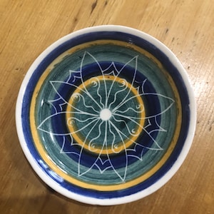 Puede incluir: Un pequeño plato de cerámica blanca con un diseño geométrico azul, verde y amarillo. El diseño presenta un círculo central con líneas radiantes y un centro blanco.