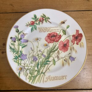 Puede incluir: Un plato decorativo blanco con borde dorado, con un diseño floral con amapolas rojas, cardos morados y margaritas. La palabra "August" está escrita en letras doradas. También hay una cita impresa.