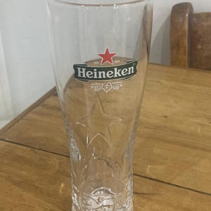 Op de afbeelding: Een helder, hoog bierglas met het Heineken-logo, met een rode ster en groene tekst. Het glas heeft een sterontwerp dat in het onderste gedeelte is ge&euml;tst en is ontworpen voor bier. Het glas is leeg en staat op een houten oppervlak.