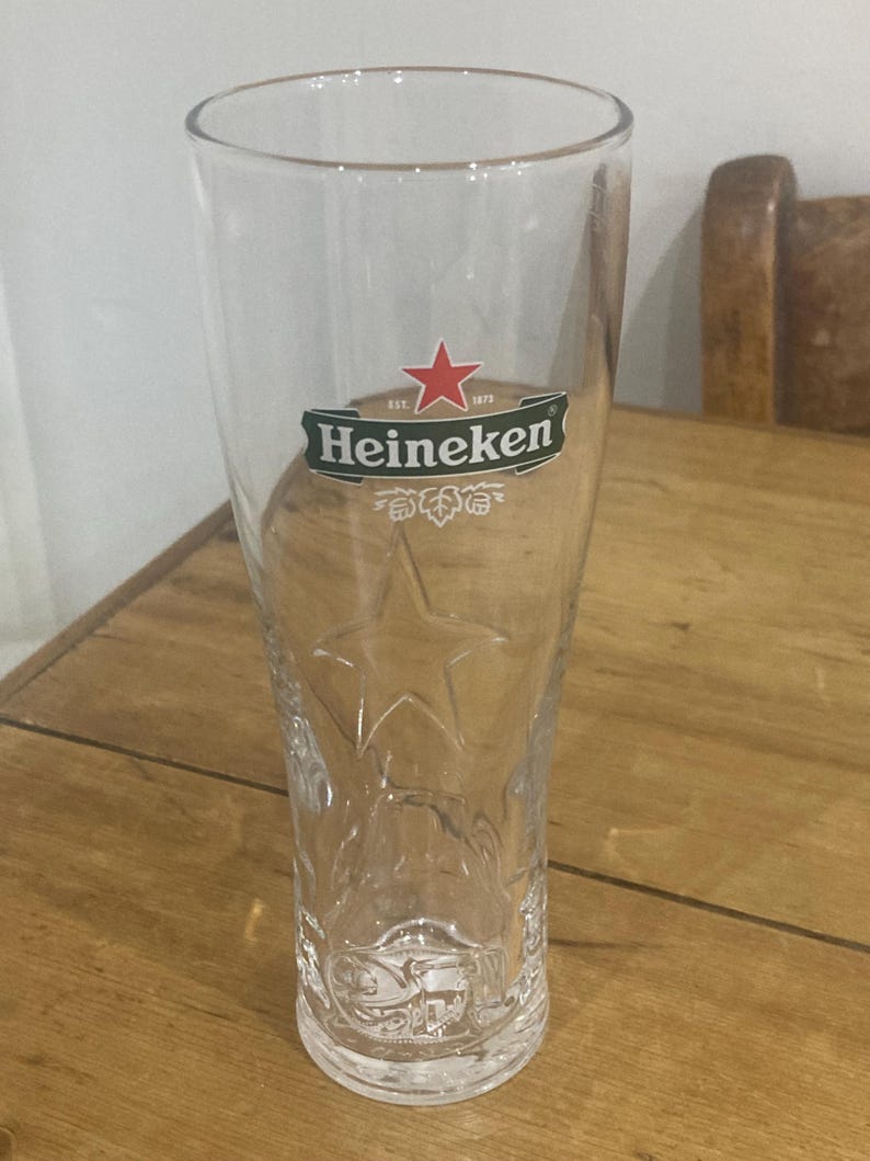 Op de afbeelding: Een helder, hoog bierglas met het Heineken-logo, met een rode ster en groene tekst. Het glas heeft een sterontwerp dat in het onderste gedeelte is ge&euml;tst en is ontworpen voor bier. Het glas is leeg en staat op een houten oppervlak.