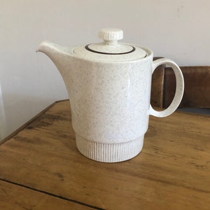 May include: A white ceramic teapot with a speckled finish and a brown stripe around the lid. The teapot has a rounded body and a handle on the side.