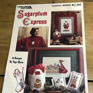 Puede incluir: Un folleto de manualidades titulado "Sugarplum Express" de Leisure Arts, con diseños de punto de cruz. La portada muestra a una mujer con un suéter rojo, junto con imágenes de proyectos de punto de cruz con temática navideña terminados, incluyendo calcetines, almohadas y arte enmarcado.