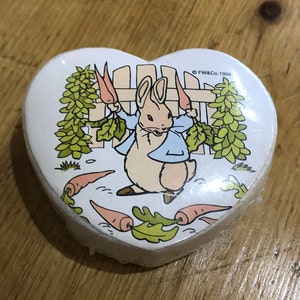 May include: A white heart-shaped box featuring a Peter Rabbit illustration. The rabbit is holding two carrots and is surrounded by green leaves and a white picket fence. The text "FW&Co. 1999" is printed on the box.