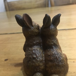 May include: Two brown ceramic rabbits sitting back to back. The rabbits have long ears and are detailed with fur texture.