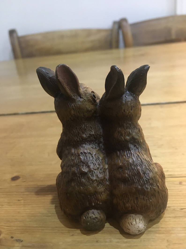 May include: Two brown ceramic rabbits sitting back to back. The rabbits have long ears and are detailed with fur texture.