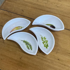 May include: Four white ceramic appetizer dishes with a leaf-shaped design. Each dish features a different botanical illustration: a chilli pepper, an olive, rosemary, and parsley. The dishes are arranged on a wooden surface.