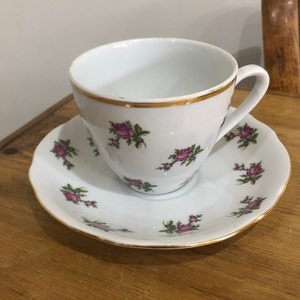 Puede incluir: Un juego de taza y platillo blanco con ribete dorado y un patrón de rosas rosas.