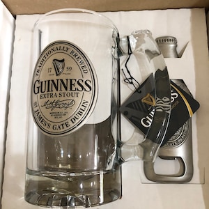May include: A silver Guinness beer mug with a black and white logo that reads "Traditionally Brewed 17 Trade Mark 59 Guinness Extra Stout Arthur's Brewed in Ireland by Guinness U.C. Ireland St. James's Gate Dublin". The mug is in a white box with a silver bottle opener.