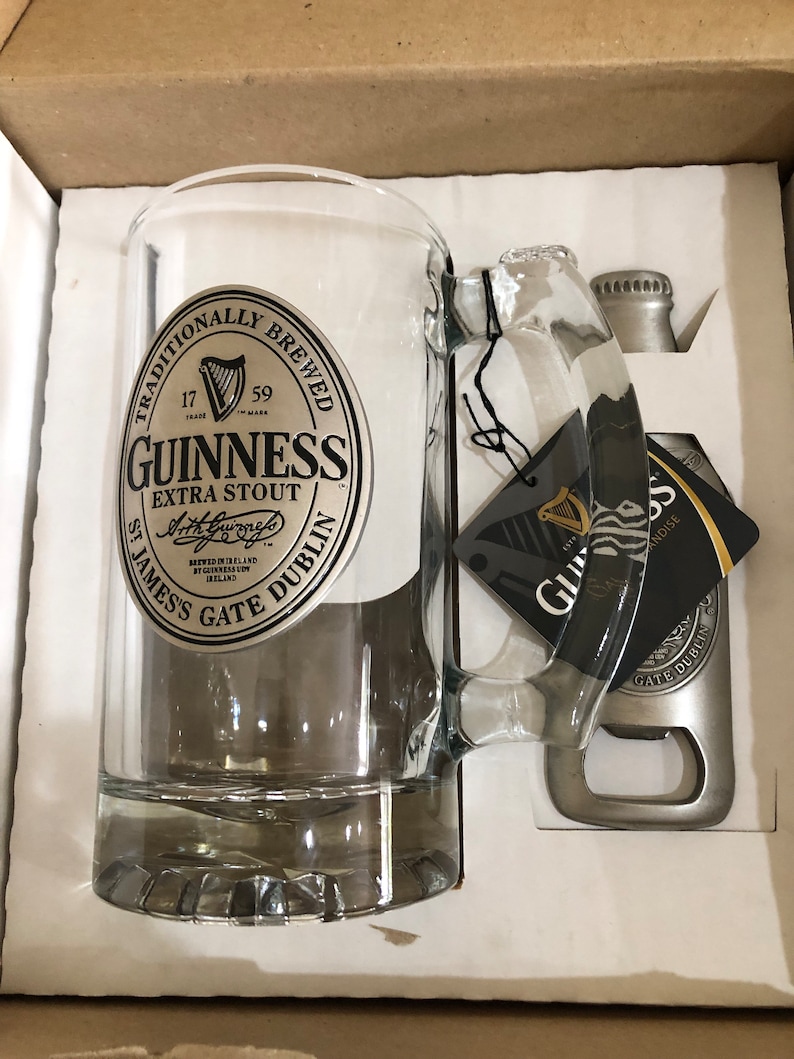 May include: A silver Guinness beer mug with a black and white logo that reads "Traditionally Brewed 17 Trade Mark 59 Guinness Extra Stout Arthur's Brewed in Ireland by Guinness U.C. Ireland St. James's Gate Dublin". The mug is in a white box with a silver bottle opener.