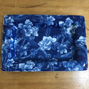 Könnte beinhalten: Ein rechteckiger Aschenbecher aus blau-weißem Keramik mit einem Blumenmuster. Der Aschenbecher hat einen leicht erhöhten Rand.