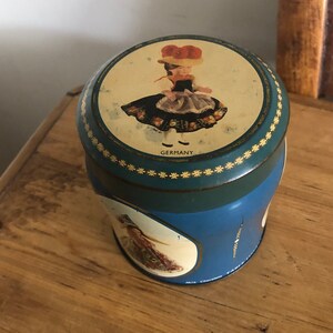 Puede incluir: Una lata azul y verde con una imagen de una niña con un vestido tradicional alemán. La tapa tiene un borde dorado con un patrón floral blanco. El texto "GERMANY" está impreso en la tapa.