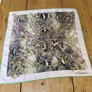 May include: Square silk scarf with a snakeskin pattern in shades of gray and tan. The scarf features a design of gold chains, brown straps, and purple accents. The border is light pink with a green trim, and the scarf has the word "Codello" in the corner.