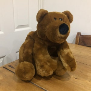 May include: A brown plush teddy bear sitting on a wooden surface. The bear has black eyes and a black nose.