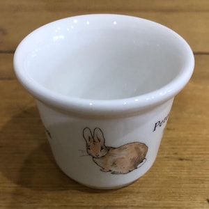 Puede incluir: Un huevo de cerámica blanca con una ilustración de conejo marrón y el texto "Peter Rabbit" en el lateral.