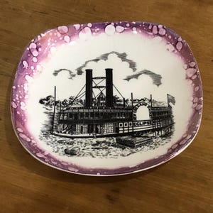 Op de afbeelding: Een klein, vierkant decoratief bord met een witte kern en een paarse rand. Het bord heeft een zwart-wit illustratie van een raderstoomboot met de tekst "New Orleans Eclipse".