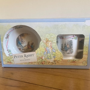 May include: A Peter Rabbit by Wedgwood ceramic bowl and mug set in a light blue and cream cardboard display box. The box features illustrations of Peter Rabbit in a garden, with the text Peter Rabbit by Wedgwood printed on the front.