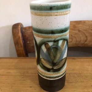 以下が含まれることがあります： 濃い茶色のベースと、白、緑、タンの斑点模様の上部を備えた、背の高い円筒形のセラミック花瓶。花瓶は、茶色、緑、青の色合いの様式化された花の模様が特徴です。花瓶の高さは約20cmです。