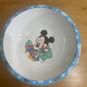 Puede incluir: Cuenco de cerámica blanca con borde azul con dibujos animados de nubes, coches y aviones. El centro del cuenco tiene un gráfico de Mickey Mouse sosteniendo un tren de juguete. El logotipo de Disney está impreso.