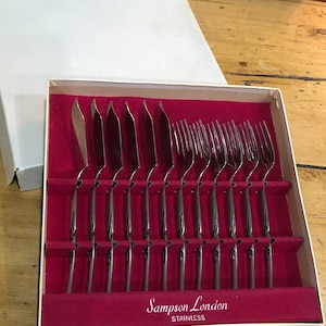 Puede incluir: Un juego de cubiertos de plata en una caja blanca con forro de terciopelo rojo. Los cubiertos están dispuestos en filas e incluyen cuchillos, tenedores y cucharas. La caja tiene las palabras "Sampson London STAINLESS" impresas en la parte inferior.