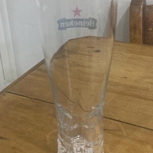 Op de afbeelding: Een helder, leeg Heineken pintglas met een sterlogo en de merknaam. Het glas is lang en slank, met een licht uitlopende bovenkant en decoratieve details aan de basis. Het staat op een houten oppervlak. Ongeveer 20 cm hoog.