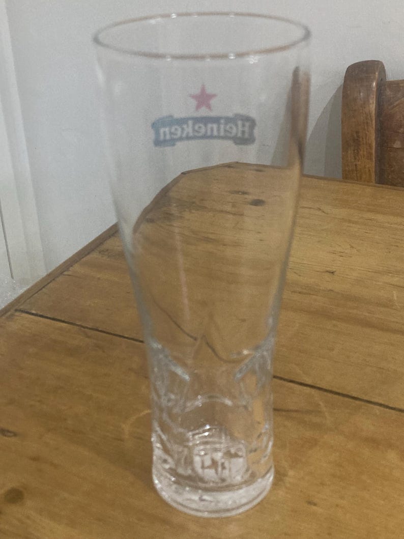 Op de afbeelding: Een helder, leeg Heineken pintglas met een sterlogo en de merknaam. Het glas is lang en slank, met een licht uitlopende bovenkant en decoratieve details aan de basis. Het staat op een houten oppervlak. Ongeveer 20 cm hoog.
