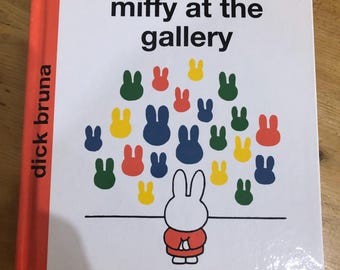 May include: A children's book titled "Miffy at the gallery" by Dick Bruna. The cover features a white bunny with red clothing standing in front of a white wall with a colorful display of bunny silhouettes.
