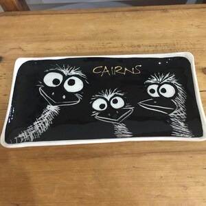 Könnte beinhalten: Rechteckige Servierplatte aus schwarzer Keramik mit weißem Rand. Sie zeigt drei Cartoon-Emu-Gesichter und den goldenen Text "CAIRNS". Die Emus sind weiß mit schwarzen Umrissen.