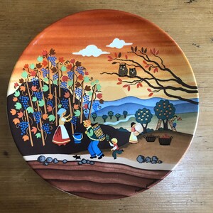 Può includere: Piatto decorativo in ceramica raffigurante una scena di vendemmia. Il piatto presenta una calda tavolozza di colori con un cielo arancione, uva blu e terreno marrone. Due gufi appollaiati su un ramo.