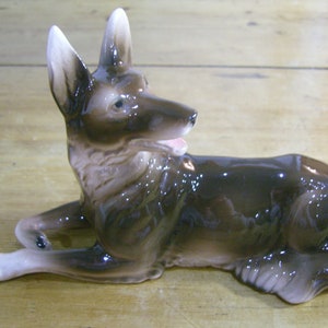 Puede incluir: Una figurita de cerámica marrón y blanca de un perro pastor alemán acostado. El perro tiene un acabado brillante y mira hacia la derecha.