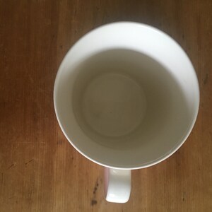 Peut inclure: Vue de dessus d'une tasse en c&eacute;ramique blanche avec une anse incurv&eacute;e. L'int&eacute;rieur de la tasse est visible, et la poign&eacute;e a un accent rose. La tasse est pos&eacute;e sur une surface en bois, soulignant le contraste.