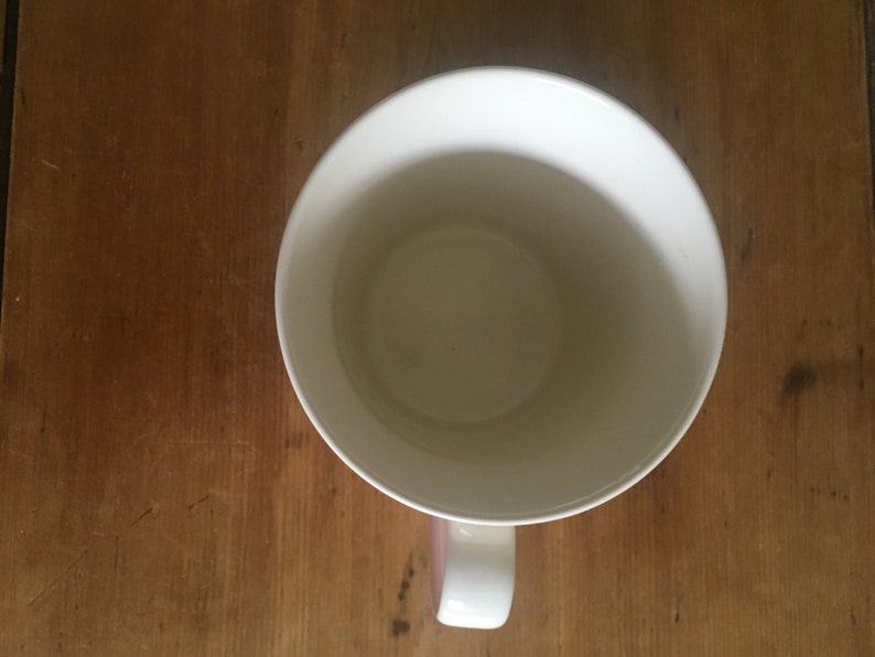 Peut inclure: Vue de dessus d'une tasse en c&eacute;ramique blanche avec une anse incurv&eacute;e. L'int&eacute;rieur de la tasse est visible, et la poign&eacute;e a un accent rose. La tasse est pos&eacute;e sur une surface en bois, soulignant le contraste.