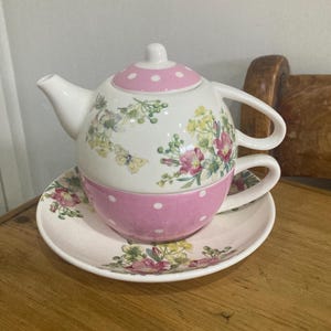 Puede incluir: Un juego de té floral blanco y rosa con tetera, taza y platillo. La tapa de la tetera y la taza tienen un diseño de lunares rosas. El juego está decorado con ilustraciones florales y de mariposas.