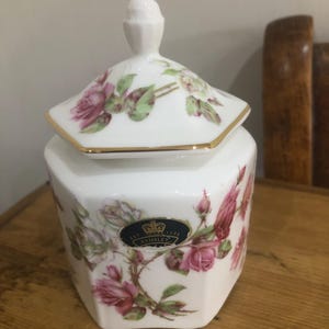May include: White porcelain jar with a lid, adorned with pink roses and green foliage. The jar features a gold trim and a small, dark blue label with gold lettering. The lid is topped with a small knob.