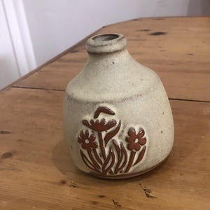 May include: A small, beige ceramic vase with a rounded body and a narrow neck. The vase features a brown floral design on the front. The vase is sitting on a wooden surface.