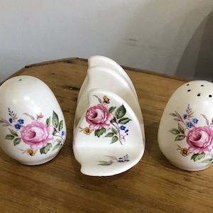 May include: A set of three white ceramic salt and pepper shakers with a floral design. The shakers are shaped like eggs and have a floral pattern with pink roses, green leaves, and blue flowers. The set also includes a white ceramic napkin holder with a floral design.