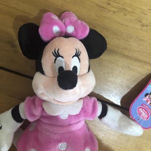 May include: Pink plush Minnie Mouse toy wearing a pink polka dot dress and a pink bow with white polka dots. The toy has black eyes, a black nose, and a white face. The toy is sitting on a wooden surface. The tag says "Disney Store Exclusive".